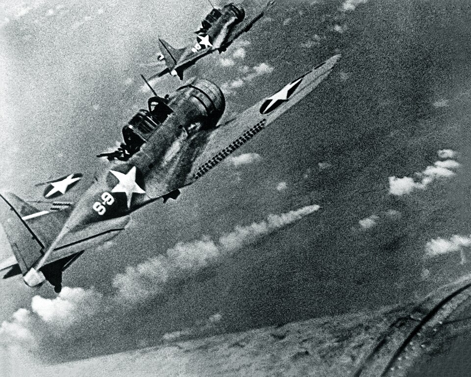 SBD Dauntless dive bombers fly in formation above the burning Japanese heavy cruiser Mikuma on June 6, 1942, bombs visible beneath their wings.