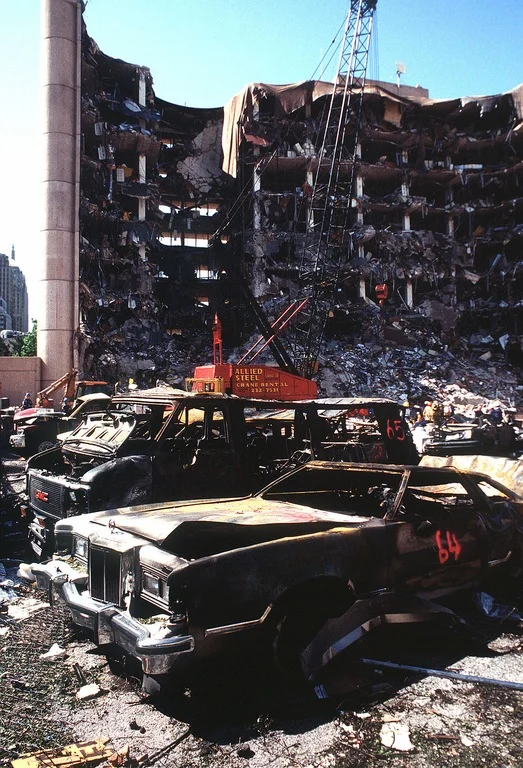 Ground-level view of the Alfred P. Murrah Federal Building in the days following the April 19, 1995 bombing, showing incinerated vehicles in the foreground and the gutted structure behind